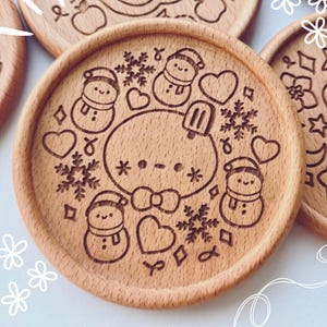 Puede incluir: Un conjunto de posavasos redondos de madera con diseños grabados. Un posavasos presenta un muñeco de nieve, corazones, copos de nieve y un personaje con un lazo. Otros posavasos tienen diferentes patrones grabados. Los posavasos son de color marrón claro.