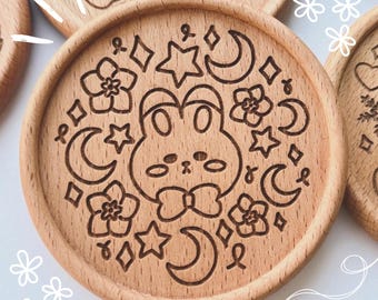 May include: Wooden coasters with engraved designs. One coaster features a rabbit face surrounded by stars, moons, and flowers. Other coasters have similar engraved patterns. The coasters are made of light-colored wood.