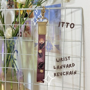 Peut inclure: Un cordon de poignet marron et doré avec un porte-clés. Le cordon présente une image imprimée d'un personnage du jeu vidéo Genshin Impact. Le texte "ITTO" et "WRIST LANYARD + KEYCHAIN" sont visibles sur l'image.