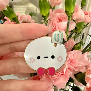 May include: A white enamel pin with a gold border, featuring a cartoon character with a popsicle, bow tie, and snowflake accents. The pin is held in front of a bouquet of pink carnations.