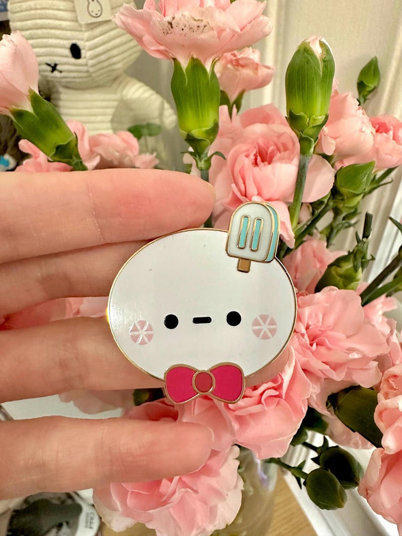 May include: A white enamel pin with a gold border, featuring a cartoon character with a popsicle, bow tie, and snowflake accents. The pin is held in front of a bouquet of pink carnations.