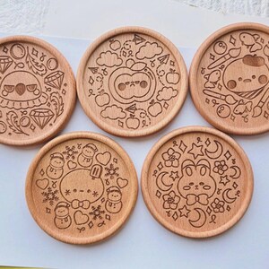 May include: Set of five round wooden coasters with engraved designs. Each coaster features a different whimsical illustration, including a diamond-themed design, an apple with a face, a painter character, snowmen, and a bunny with moons and stars.