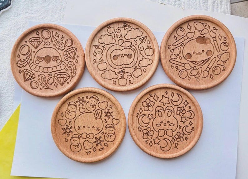 May include: Set of five round wooden coasters with engraved designs. Each coaster features a different whimsical illustration, including a diamond-themed design, an apple with a face, a painter character, snowmen, and a bunny with moons and stars.