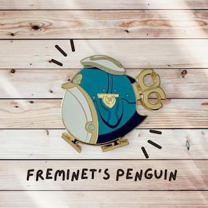 May include: A gold-colored penguin pin with a blue and white design. The penguin is wearing a hat and has a key-shaped tail. The pin is on a wooden background with the text "FREMINET'S PENGUIN".