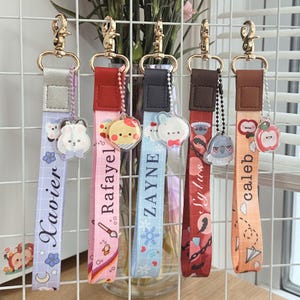 May include: A collection of personalized lanyards with gold-colored clasps. Each lanyard features a different color strap with a name printed on it, along with a charm. The charms include a bunny, a chick, a koala, and an apple.