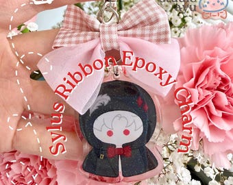 Pode incluir: Um pingente de epóxi com um personagem em uma capa escura com capuz, olhos vermelhos e gravata borboleta vermelha, finalizado com um laço de tecido xadrez rosa e tecido transparente. Inclui um fecho de metal prateado e o texto Sylus Ribbon Epoxy Charm.
