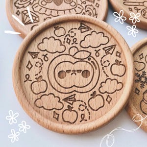 May include: Wooden coasters with engraved designs. One coaster features a cartoon apple with a grumpy face, surrounded by clouds, paper airplanes, and apples. Other coasters have similar engraved illustrations.