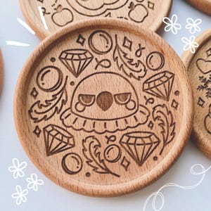 Peut inclure: Sous-verres en bois avec des motifs gravés. L'un des dessous de verre présente un personnage central à l'expression renfrognée, entouré de diamants, de bulles et d'éléments décoratifs. Les dessous de verre sont en bois clair.