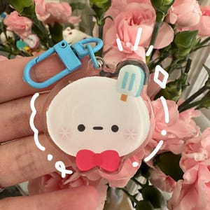 Puede incluir: Un llavero acrílico transparente con un personaje de dibujos animados con una cara blanca, ojos negros y una pajarita roja. El llavero tiene un cierre azul y un diseño de helado. El fondo incluye claveles rosas.