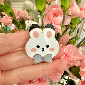 May include: A white bunny pin with black ears, pink cheeks, and a gray bow tie. The pin is made of metal and enamel. The bunny pin is a cute accessory.