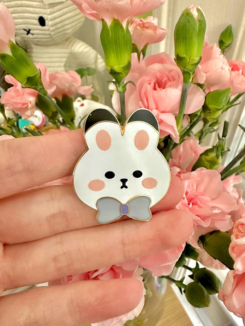 May include: A white bunny pin with black ears, pink cheeks, and a gray bow tie. The pin is made of metal and enamel. The bunny pin is a cute accessory.