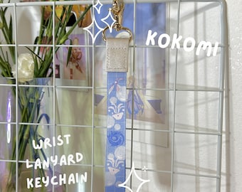 Pode incluir: Um chaveiro de cordão de pulso azul e branco com padrão com a palavra "KOKOMI" escrita em branco sobre um fundo azul. O chaveiro está pendurado em uma grade de metal branca.