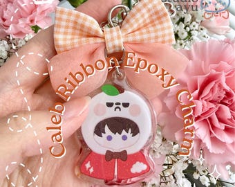 Pode incluir: Um pingente de resina epóxi com um personagem de desenho animado em um capuz branco e capa vermelha, coberto com um laço de tecido xadrez laranja. O pingente exibe o texto Caleb Ribbon Epoxy Charm e Mascot Fit com uma folha verde.