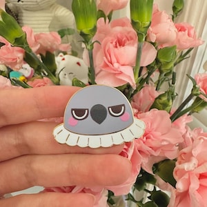 May include: A gray enamel pin with a grumpy bird design. The pin features a white ruffled collar, pink cheeks, and a black beak. The pin is held in front of a bouquet of pink carnations.
