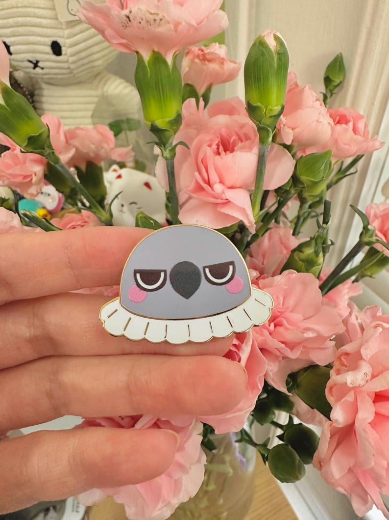 May include: A gray enamel pin with a grumpy bird design. The pin features a white ruffled collar, pink cheeks, and a black beak. The pin is held in front of a bouquet of pink carnations.