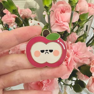 May include: A pink enamel pin in the shape of an apple with a grumpy face. The pin has a gold border, a green leaf, and a brown stem. The background features pink carnations.
