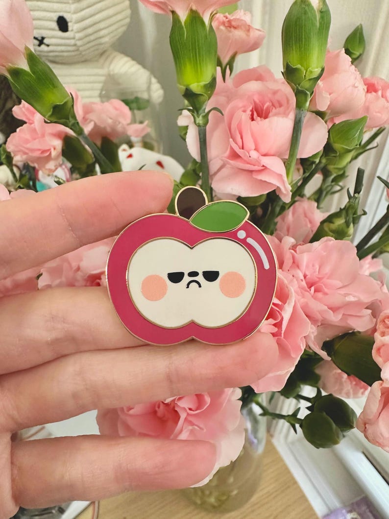 May include: A pink enamel pin in the shape of an apple with a grumpy face. The pin has a gold border, a green leaf, and a brown stem. The background features pink carnations.