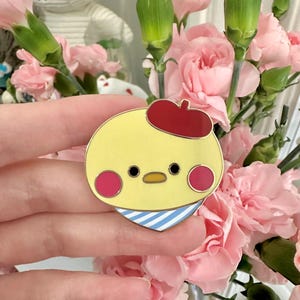 May include: A yellow enamel pin featuring a cartoon chick wearing a red beret and a blue and white striped scarf. The chick has pink cheeks and black eyes. The pin is held in front of a bouquet of pink carnations.