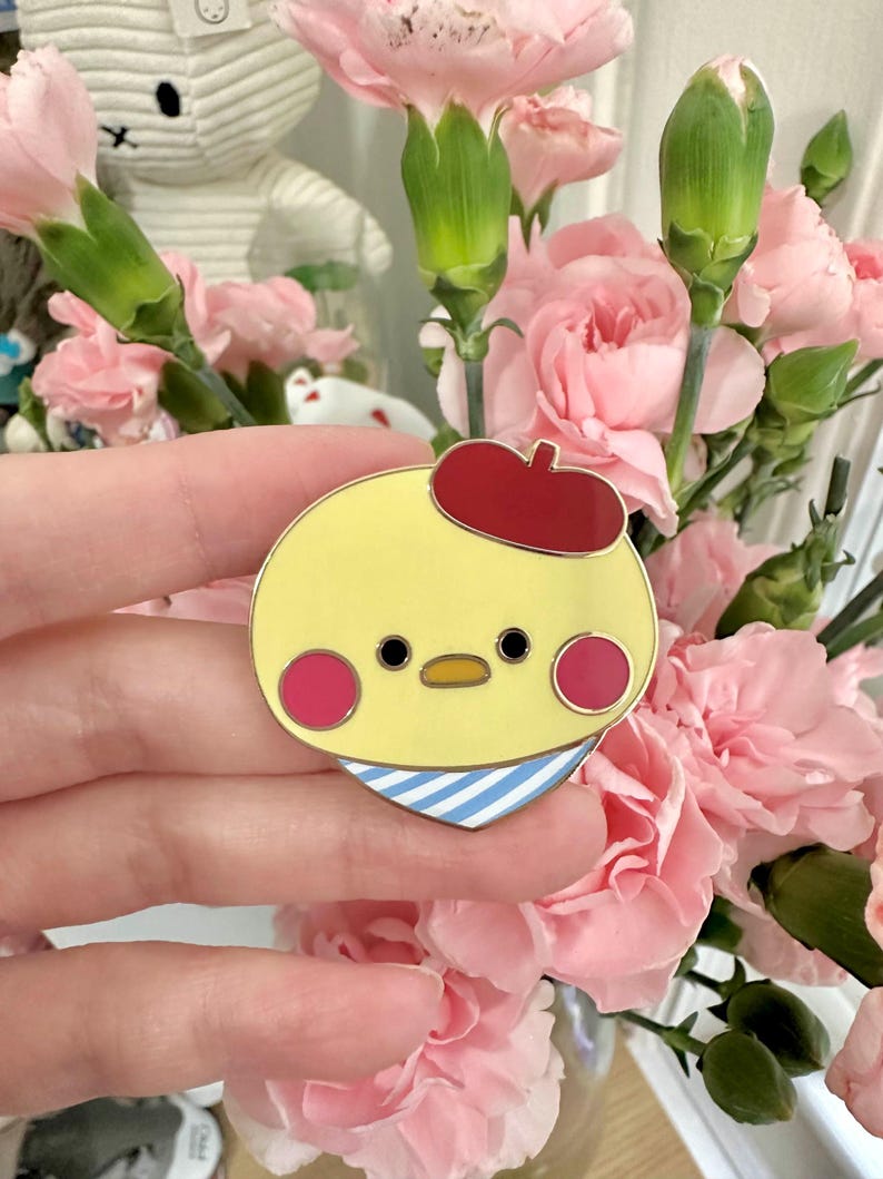 May include: A yellow enamel pin featuring a cartoon chick wearing a red beret and a blue and white striped scarf. The chick has pink cheeks and black eyes. The pin is held in front of a bouquet of pink carnations.