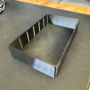 May include: A black plastic storage tray with dividers. The tray is empty and has a smooth, glossy finish.