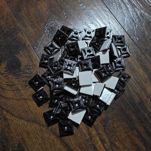 May include: A pile of black square cable tie mounts with a central hole, and white adhesive pads. The mounts are made of plastic and are designed to secure cables. The background is a dark wood surface.