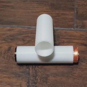 May include: Two white cylindrical battery holders, one upright and one horizontal, with a copper-colored end visible on the horizontal battery. The holders are on a brown wooden surface.