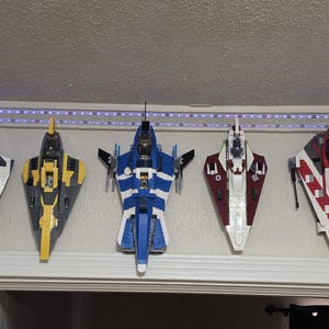 May include: Five colorful toy spaceships are mounted on a wall. The ships are various colors, including black, yellow, blue, red, and white. They are arranged in a row, with a light strip above them.