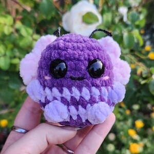 May include: A small, crocheted plush toy in the shape of a purple and pink creature with large black eyes. The toy has a smiling face and a textured, fuzzy surface.