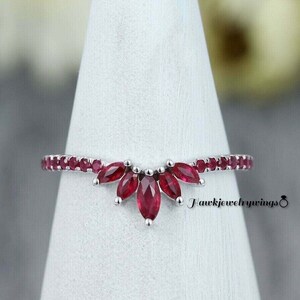 May include: A delicate silver ring with a curved band of small red gemstones and a larger cluster of five red marquise-shaped gemstones in the center. The ring is displayed on a white cone.
