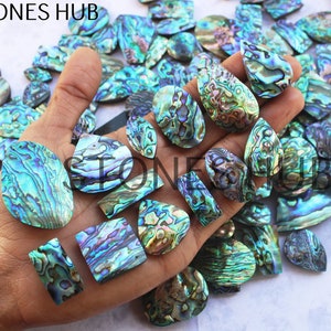Pode incluir: Uma coleção de cabochões de concha de abalone iridescente em várias formas e tamanhos. Os cabochões são uma mistura de tons verdes, azuis e roxos.