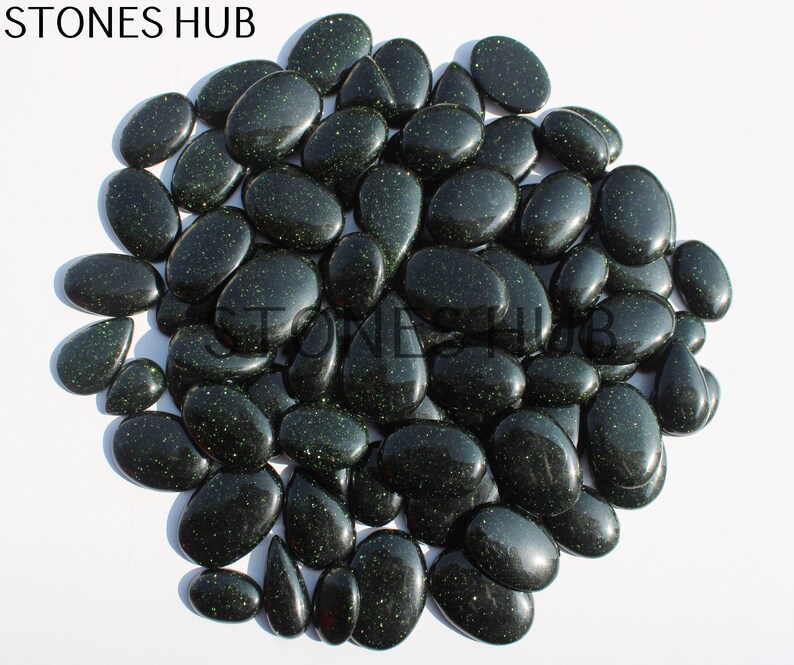 May include: A large group of dark green, oval-shaped stones with a glittery finish. The stones are arranged in a circular pattern on a white background.