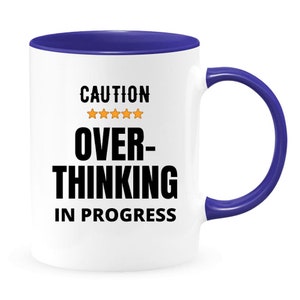 May include: White ceramic coffee mug with a blue rim and handle. The mug has the text "CAUTION" with five stars and the text "OVER-THINKING IN PROGRESS" printed on it.