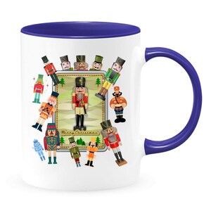 May include: White ceramic mug with a blue rim and handle. The mug features a colorful illustration of nutcracker figurines surrounding a larger nutcracker with the text "Merry Christmas".