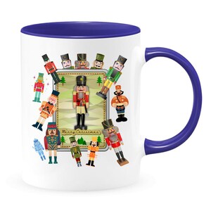 May include: White ceramic mug with a blue rim and handle. The mug features a colorful illustration of nutcracker figurines with the text "Merry Christmas".