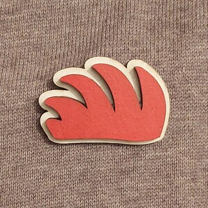 Puede incluir: Un pin de madera rojo con un diseño de llama estilizado. El pin está hecho de madera y tiene un contorno blanco.