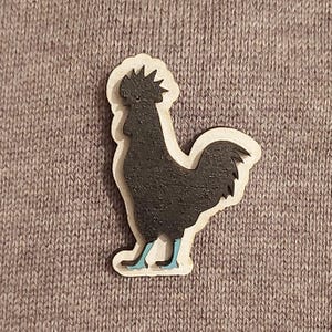 Puede incluir: Un recorte de madera de un gallo negro con patas azules. El gallo está mirando hacia la derecha y tiene una cresta y barbillas grandes.