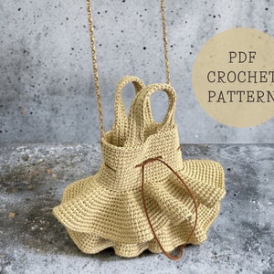 Può includere: Una borsa all'uncinetto a forma di vestito con un cinturino marrone. La borsa ha un fondo a balze ed è appesa a una catena dorata. Il testo "PDF CROCHET PATTERN" è in un cerchio sul lato destro dell'immagine.