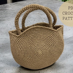 Puede incluir: Un bolso de mano tejido a crochet con fondo redondo y dos asas. El bolso está hecho de hilo marrón natural.