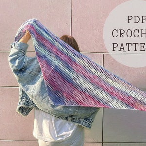 Peut inclure: Une personne portant une veste en jean tient un châle crocheté avec un motif rayé multicolore. Le châle est de couleur bleu clair, rose et blanc. Le texte "PDF CROCHET PATTERN" est dans un cercle dans le coin supérieur droit de l'image.