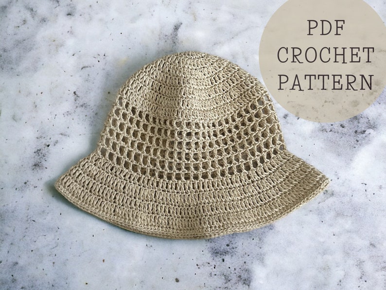 May include: A crocheted beige bucket hat with a lacy pattern. The hat is on a white marble surface. The text "PDF CROCHET PATTERN" is in a circle on the right side of the image.