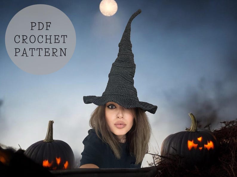 May include: A black crochet witch hat with a tall, pointed crown. The hat is worn by a person with long brown hair. The background features a blurry image of a moon and two jack-o'-lanterns.