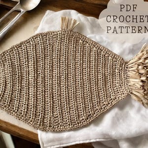 May include: A beige, fish-shaped crochet pattern, with a textured surface and a fringed tail. The text "PDF CROCHET PATTERN" is in a white cloud-shaped text box. The crochet item is on a white cloth.