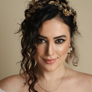 Może przedstawiać: Kobieta z długimi, ciemnymi, kręconymi włosami, kt&oacute;ra nosi złotą koronę z liści i złoty naszyjnik. Ma na sobie białą bluzkę z odkrytymi ramionami.