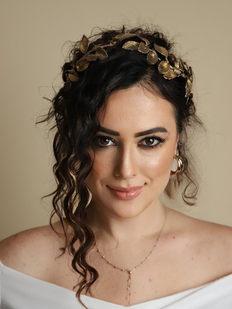 Może przedstawiać: Kobieta z długimi, ciemnymi, kręconymi włosami, kt&oacute;ra nosi złotą koronę z liści i złoty naszyjnik. Ma na sobie białą bluzkę z odkrytymi ramionami.