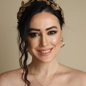 Może przedstawiać: Kobieta o ciemnych włosach w złotej opasce do włos&oacute;w z liści i złotym naszyjniku. Uśmiecha się i patrzy w obiektyw.