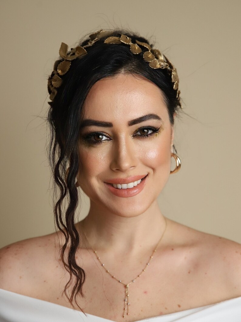 Może przedstawiać: Kobieta o ciemnych włosach w złotej opasce do włos&oacute;w z liści i złotym naszyjniku. Uśmiecha się i patrzy w obiektyw.