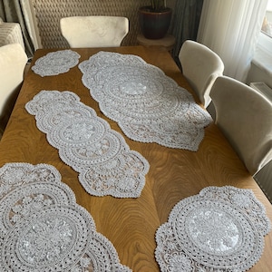 May include: A set of four silver lace table runners with intricate floral designs. The runners are arranged on a wooden table with chairs.