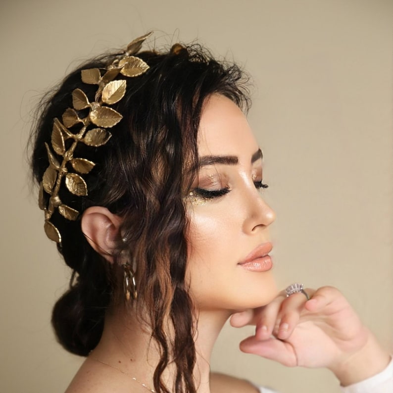 Może przedstawiać: Kobieta z ciemnobrązowymi, falującymi włosami, kt&oacute;ra nosi złotą koronę z liści. Ma naturalny makijaż z  złotym brokatem na powiekach.