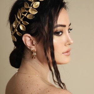 Może przedstawiać: Kobieta z długimi, ciemnymi włosami, kt&oacute;ra nosi złotą opaskę na głowę w kształcie korony z liści. Ma na sobie białą bluzkę bez ramiączek i złote kolczyki koła.