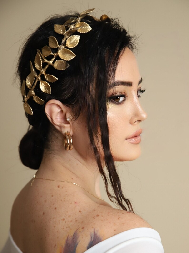 Może przedstawiać: Kobieta z długimi, ciemnymi włosami, kt&oacute;ra nosi złotą opaskę na głowę w kształcie korony z liści. Ma na sobie białą bluzkę bez ramiączek i złote kolczyki koła.